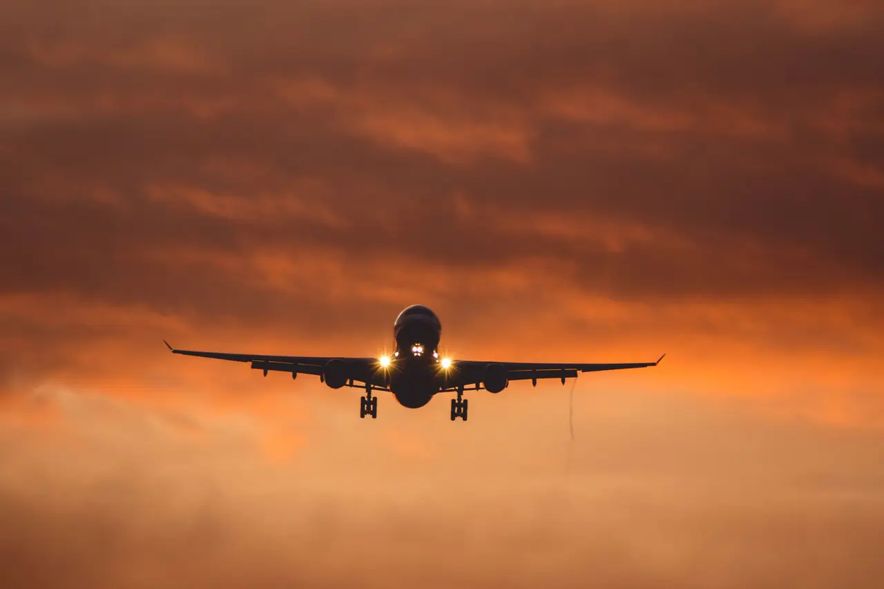 A twin-engine jet with landing lights on and gear down, silhouetted against an orange dusk sky