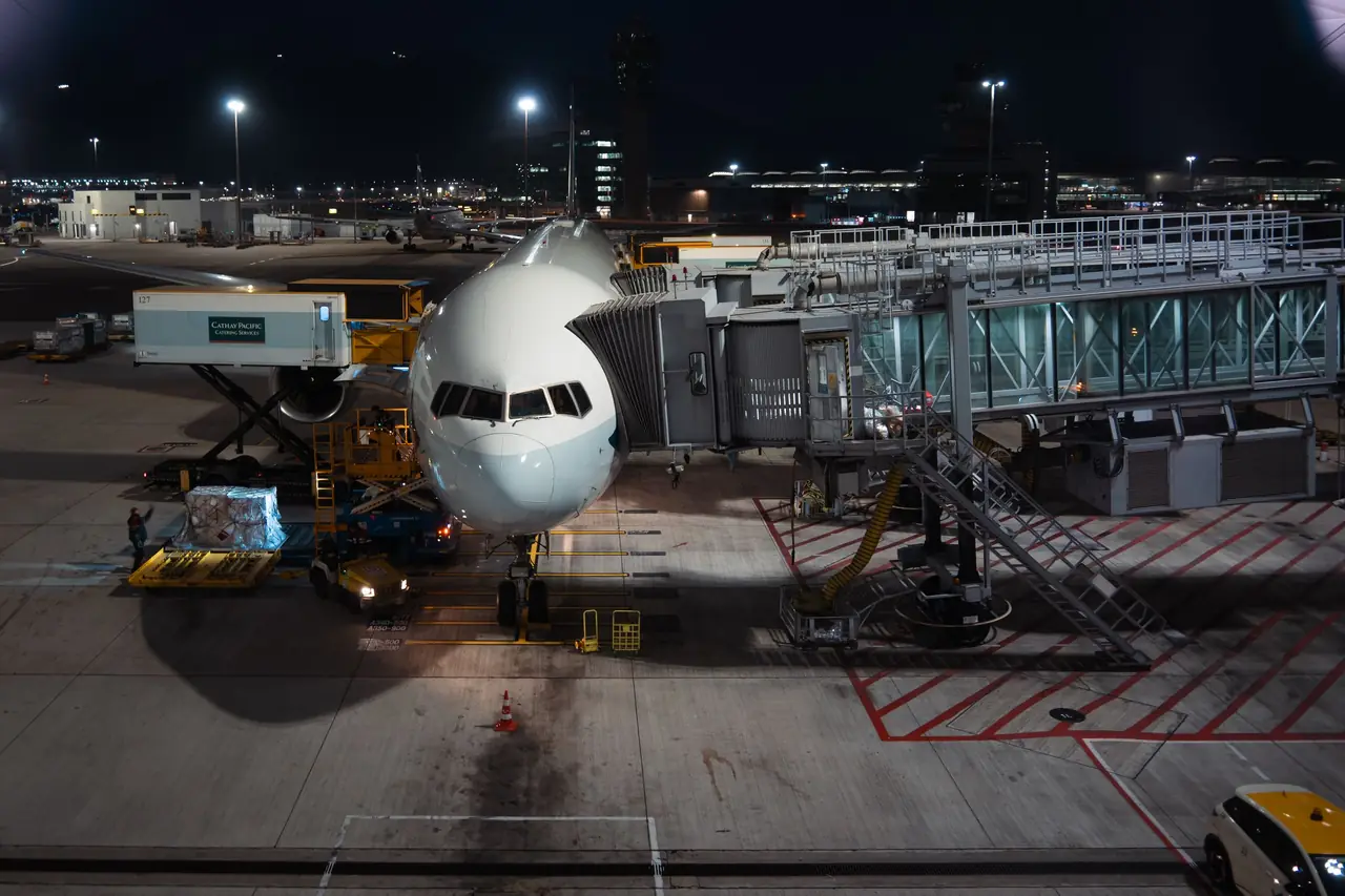 A widebody jet parked at a jet bridge on a floodlit apron at night while ground crews load cargo