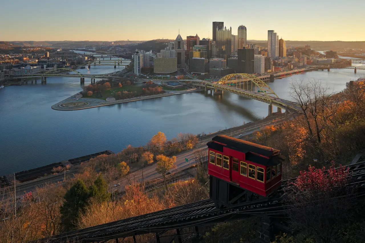 A red Duquesne Incline car climbing Mount Washington above the Point, with downtown Pittsburgh and the yellow Fort Duquesne Bridge in morning light