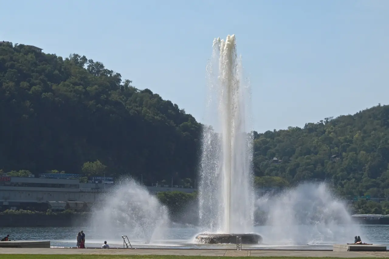 The Point State Park fountain shooting a tall column of water at the tip of downtown Pittsburgh, with wooded Mount Washington across the river